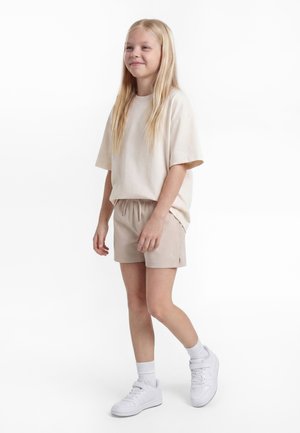 Νεαρή κοπέλα με μακριά ξανθά μαλλιά στέκεται, φοράει μπεζ μεγάλη μπλούζα, μπεζ σορτς, λευκές κάλτσες και λευκά αθλητικά παπούτσια με φόντο λευκό χρώμα.