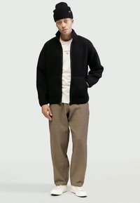 Must musta zip-up fliisjakker, millel on tekstuurne viimistlus, valge T-särgi peal, beežide pükste ja valgete tossudega.