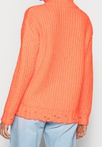Pull en maille épaisse de couleur orange vif avec un col montant et une texture côtelée. Présente une coupe ample et un détail de ourlet usé.
