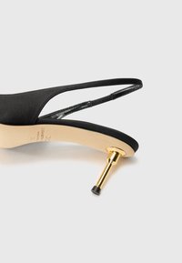Musta satiininen slingback-korkokenkä, jossa on kärjessä terävä muoto ja kultainen metallinen yksityiskohta kantapäässä. Sisältää minimalistisen muotoilun ja sulavat nauhat.