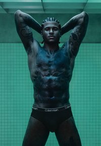 Schwarze Calvin Klein Slips getragen von einer muskulösen männlichen Figur mit geflochtenem Haar und Tattoos an beiden Armen, vor einem grünen gefliesten Hintergrund.