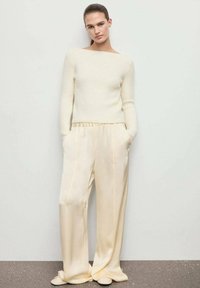 Una giovane donna è in piedi contro un muro, indossa un maglione a maglia color crema e pantaloni larghi in satin con le mani nelle tasche.