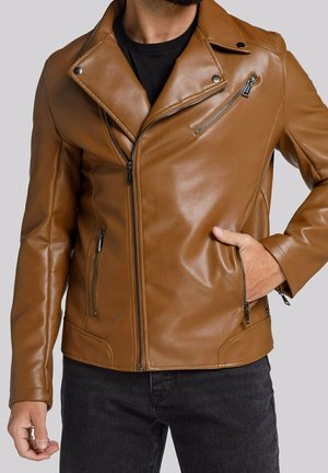 Mężczyzna w brązowej skórzanej kurtce motocyklowej z kieszeniami na zamek, na czarnej koszulce i ciemnych dżinsach, z ręką w kieszeni kurtki.