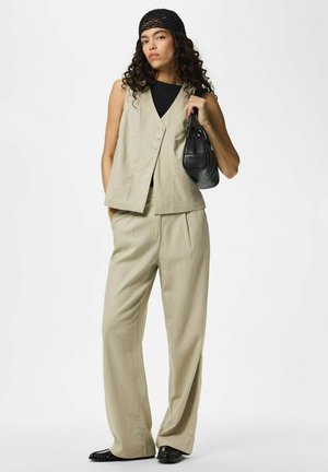 Femme aux longs cheveux bouclés portant un gilet beige et un pantalon à jambes larges, un haut noir, un foulard noir sur la tête et tenant un sac à main noir.