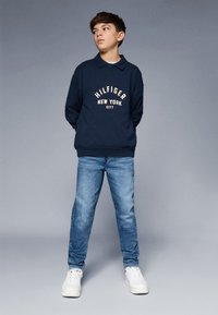 Sötétkék pulóver "HILFIGER NEW YORK CITY" grafikával, világoskék farmernadrággal és fehér sportcipővel párosítva. Laza, kényelmes fazon.