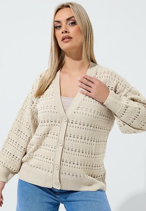 Blondynka ubrana w beżowy, otwarty sweter zapinany na guziki, na jasnej bluzce i niebieskich dżinsach, pozująca z jedną ręką przy kołnierzu.