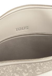 Wnętrze torby TOUS w jasnym beżowym kolorze z zamkiem błyskawicznym, charakteryzujące się gładką teksturą i wytłoczonym logo, otoczone subtelnym wzorem.