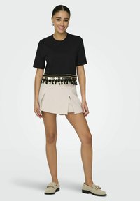 T-shirt noir court avec un ourlet décoratif en franges dorées et noires, associé à une mini-jupe plissée beige et des mocassins beiges.