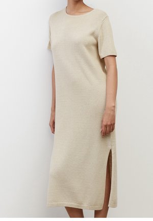 Beige strikket midi-kjole med korte ærmer og sideslids, båret af en stående person mod en ensartet lys baggrund.