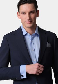 Navy blue suit jacket with subtle texture, light blue dress shirt, and patterned pocket square. Cuff with black button detail visible.
