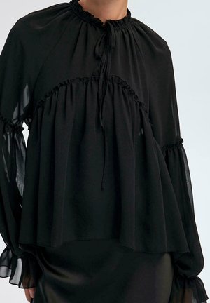 Blouse noire à manches longues avec col montant à volants, détails froncés et manches longues transparentes et fluides nouées aux poignets, portée sur une jupe noire.