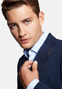 Granatowa, teksturowana marynarka z jasnoniebieską koszulą z kołnierzykiem; posiada subtelny wzór i ozdobną przypinkę na klapie; gładka karnacja i stylowe włosy.