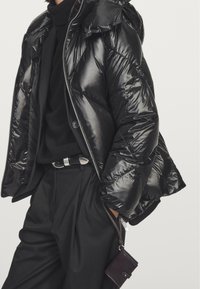 Black puffer jacket with a glossy finish, hood, and front zipper. Worn over a black sweater and high-waisted trousers, featuring a small bag.