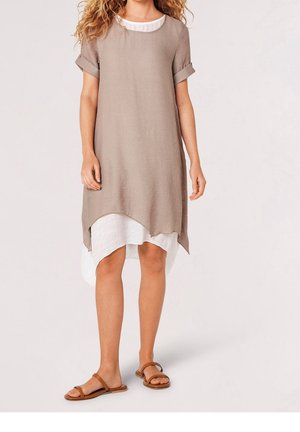 Donna che indossa un abito beige e bianco a strati, più corto davanti e più lungo dietro, con maniche corte arrotolate e sandali bassi marroni, su sfondo neutro.