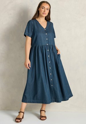 Kvinde iført en mørkeblå midi-kjole med knappelukning foran, korte ærmer og brune hælede sandaler, poserer med den ene hånd i lommen.