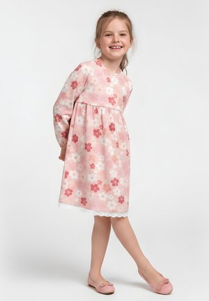 Uśmiechnięta dziewczynka w różowej sukience w kwiaty z długimi rękawami i różowych balerinach, stojąca na jednolitym jasnym tle.