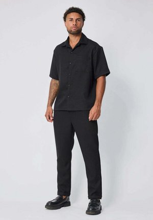 Homme aux cheveux courts et bouclés portant une chemise noire à manches courtes boutonnée, un pantalon noir et des chaussures noires, debout devant un fond uni.