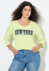 Limonkowa zielona koszula z długim rękawem i dekoltem w serek, z granatowym nadrukiem graficznym "NEW YORK" na piersi. Noszona z niebieskimi dżinsami.