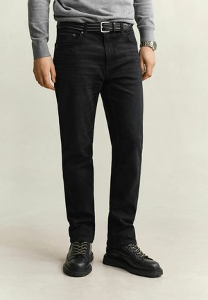 Homme portant un jean noir, une ceinture noire, des bottes noires à lacets, un pull gris et une montre-bracelet argentée, debout sur un fond neutre.