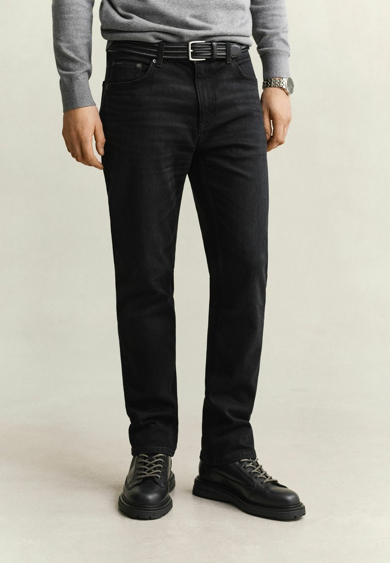 Homme portant un jean noir, une ceinture noire, des bottes noires à lacets, un pull gris et une montre-bracelet argentée, debout sur un fond neutre.