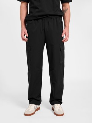 Mand iført sorte cargobukser og hvide sneakers, stående mod en ensfarvet baggrund med hænderne afslappet ned langs siderne.
