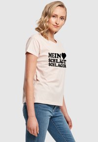 Růžové bavlněné tričko s krátkým rukávem, na kterém je černý text "MEIN ♥ SCHLÄGT SCHLAGER." Stylizováno s modrými džíny, model otočený vlevo.