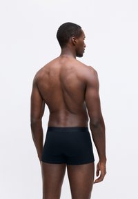 Muscular man standing barefoot facing away, wearing black fitted boxer briefs against plain white background.