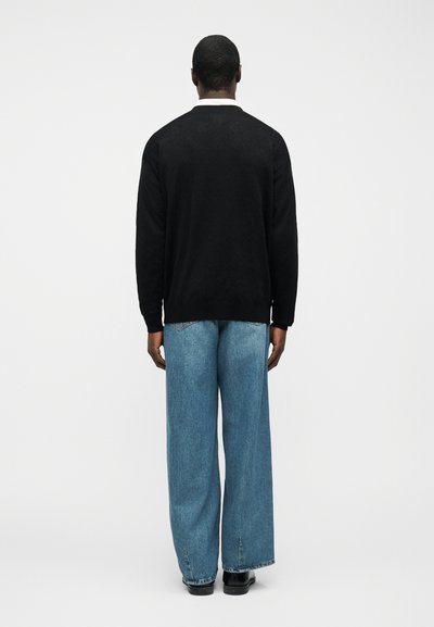 Άνδρας που φοράει μαύρο πουλόβερ, λευκό γιακά πουκάμισου, μπλε τζιν καμπάνα και μαύρα παπούτσια, στέκεται με την πλάτη στραμμένη σε απλό φόντο.