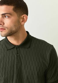 Polo de color verde oscuro con un patrón texturizado, cuello doblado y abertura de tres botones. Hecho de una tela suave, con un diseño ajustado.