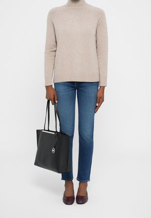 Personne portant un pull à col roulé beige, jean bleu, chaussures bordeaux, tenant un sac fourre-tout en cuir noir avec un pendentif logo circulaire.