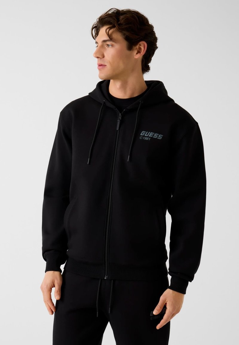 Sweat à capuche noir avec fermeture éclair, texture douce, poche avant et logo "GUESS" en turquoise. Poignets et ourlet côtelés. Design simple et décontracté.