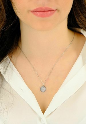 Collier en argent avec un pendentif circulaire arborant un design détaillé. La chaîne est délicate et fine, portée sur un chemisier blanc.
