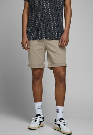 Beige Shorts mit umgeschlagenen Säumen, kombiniert mit einem schwarzen gemusterten Hemd und weißen Socken mit einem Textdesign. Schwarze und weiße Sneakers runden den Look ab.