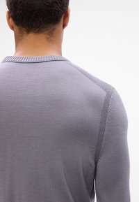 Homme vu de dos portant un pull en maille gris clair près du corps avec des détails de coutures texturées à l'épaule et à la manche.