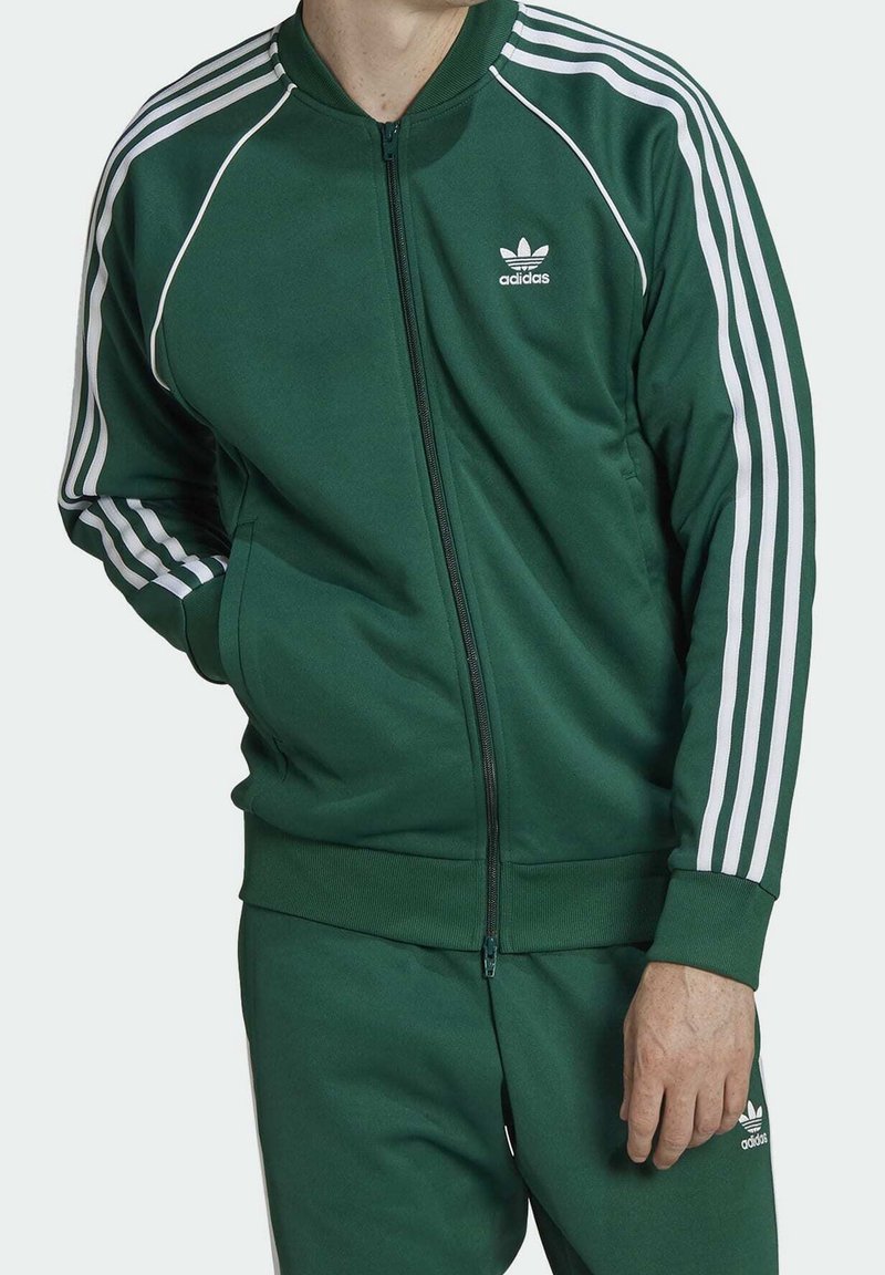 Grön zip-up tracksuitjacka med vita ränder längs ärmarna, ribbade muddarna och liten vit adidas-logotyp på bröstet.