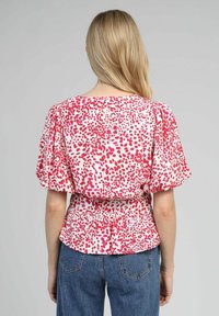 Blouse à manches courtes en tissu blanc avec un motif à taches roses et rouges, taille cintrée et coupe loose. La vue de dos montre un encolure arrondie.