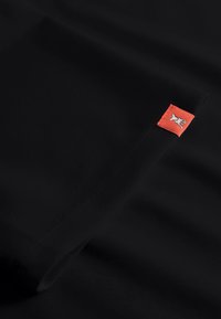 Zwarte stof met een gladde textuur, voorzien van een rood label met een witte afbeelding. De stof hangt elegant, met naadloze randen.