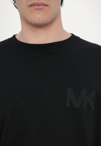 Černé bavlněné tričko s kulatým výstřihem a texturovaným, vyšívaným logem "MK" na levé straně hrudi. Hladká textura látky.