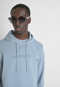 Felpa azzurra chiara con cappuccio e logo ricamato "MORATO". Abbinata a occhiali da sole quadrati con lenti scure e montatura trasparente.