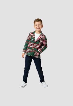 Młody chłopiec uśmiechający się, ubrany w białą koszulę, zielono-czerwony kratkowany blezer, granatowe spodnie i białe trampki, stojący z rozluźnionymi ramionami na szarym tle.