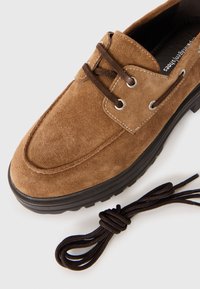 Chaussure en daim marron à bout rond, lacets marron, œillets en acier, et semelle noire texturée. Inclus un lacet marron supplémentaire.