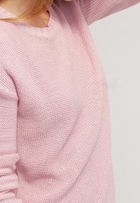 Pull tricoté rose pâle avec col en V, manches longues et motif texturé. Le matériau semble doux et léger.