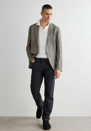 Uomo che indossa un blazer grigio a spina di pesce sopra una camicia lavorata a maglia bianca, jeans neri e sneakers nere, in piedi contro un muro bianco uniforme.