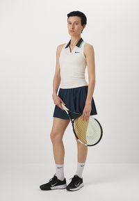 Conjunto de tenis de Nike que presenta una blusa sin mangas de color beige claro con un cuello negro y turquesa, combinada con una falda azul marino y zapatillas negras.
