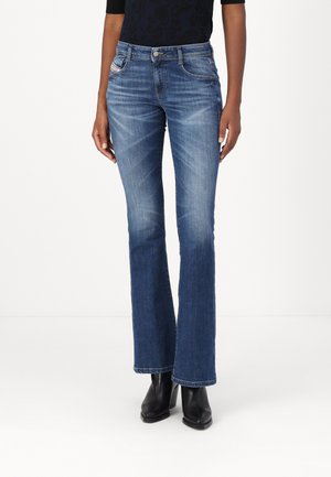 Personne portant un jean bootcut en denim bleu taille mi-haute avec légère décoloration, associé à des bottines noires à bout pointu et un haut noir.