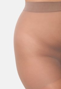 Primo piano di una persona che indossa collant beige trasparenti con una cintura elasticizzata a coste su uno sfondo chiaro.