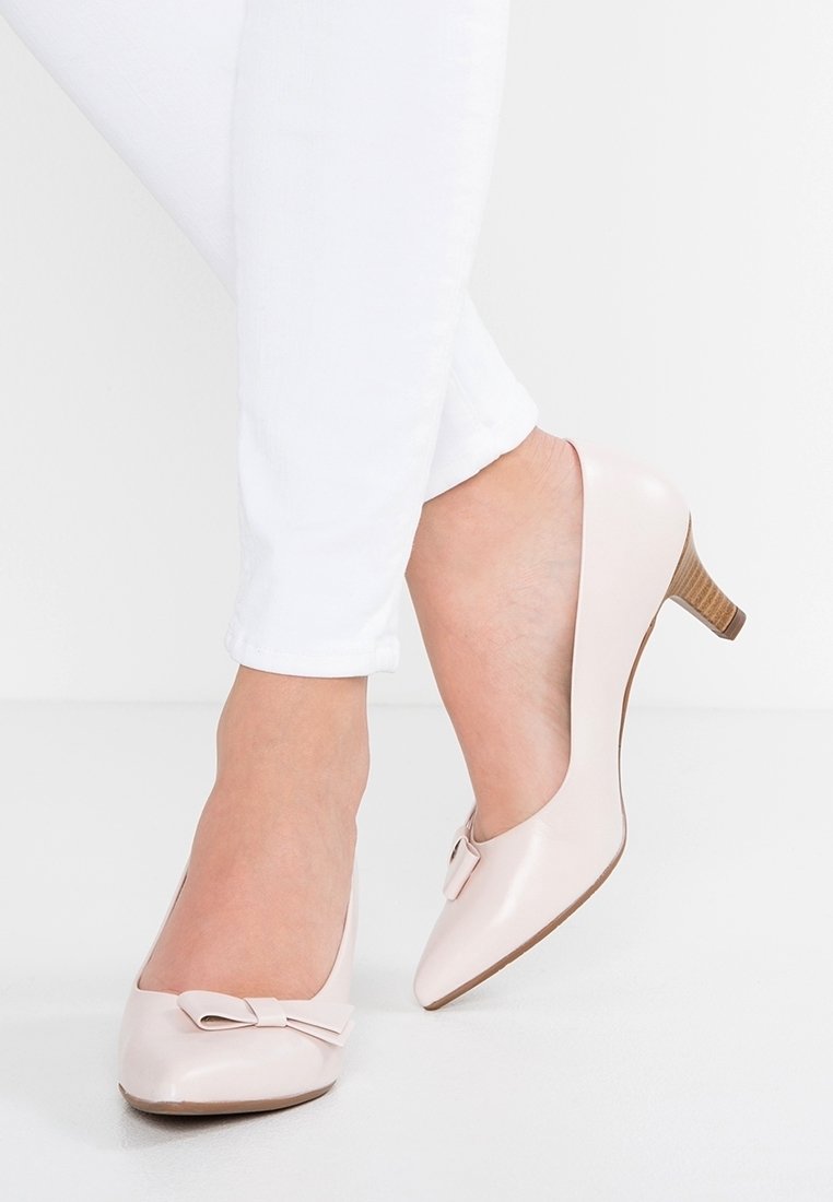 Pieds portant des chaussures à petit talon rose pâle avec un détail en nœud, assorties à un pantalon blanc slim, sur un fond blanc uni.