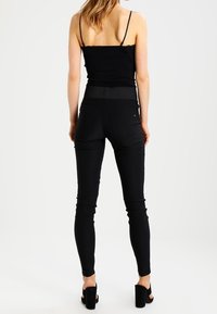 Kvinna som står vänd bort, iförd en svart ärmlös topp, högmidjade svarta skinnybyxor och svarta klacksandaler på vit bakgrund.