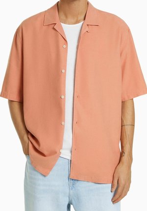 Hombre con camisa coral de manga corta abotonada sobre una camiseta blanca y vaqueros azul claro, de pie con las manos parcialmente en los bolsillos.