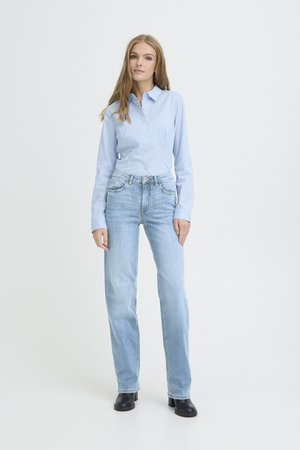 Frau steht und trägt ein hellblaues Hemd zum Knöpfen, hellblaue Jeans mit geradem Bein und schwarze Stiefeletten vor einem einfarbigen weißen Hintergrund.